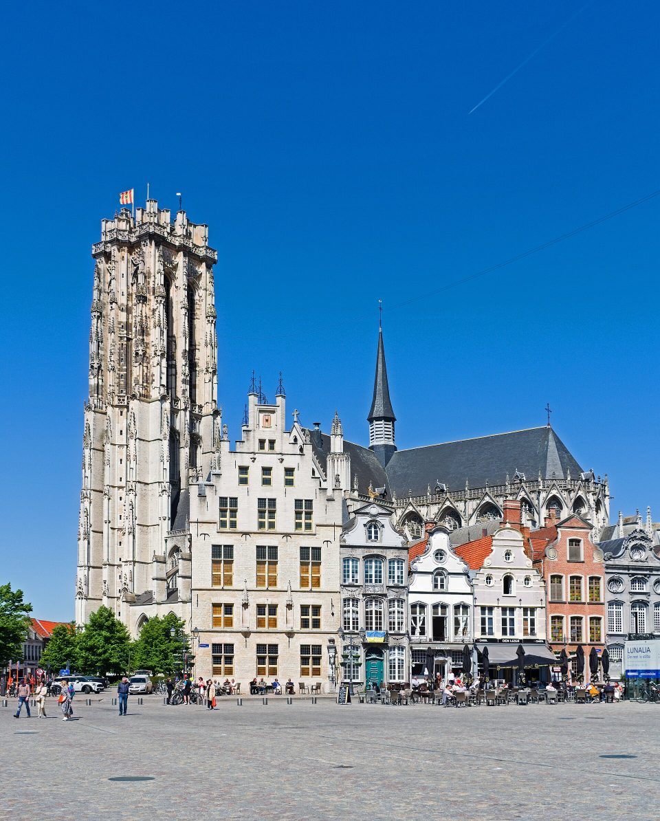 La haute tour de pierre de la cathédrale Saint-Rombaut au-dessus des façades et des terrasses de la Grand-Place de Malines, sous un ciel d’un bleu limpide