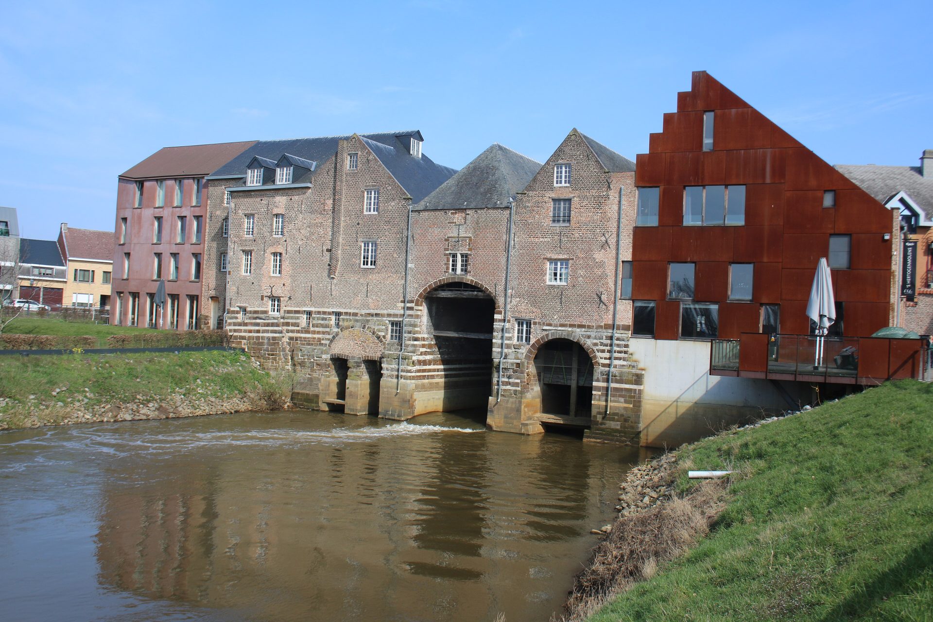 Les 's Hertogenmolens à Aarschot : d’anciens bâtiments de moulin en brique, avec des arches voûtées au-dessus du Démer et, à droite, une extension récente en acier couleur rouille, sous un ciel bleu