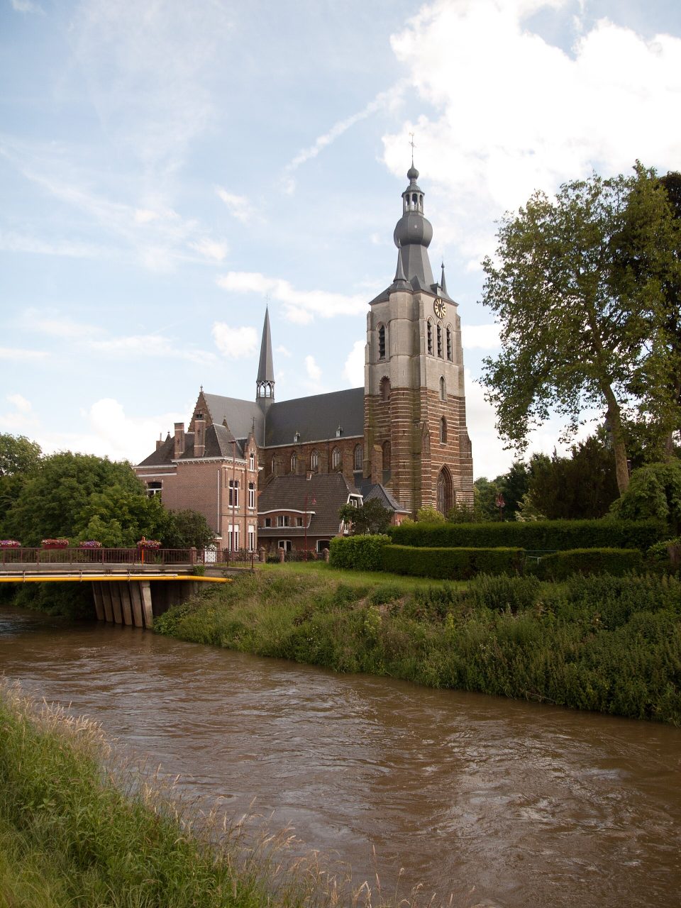 L’église Notre-Dame d’Aarschot et sa haute tour rayée, vue depuis l’autre rive du Démer, avec un pont à gauche et des arbres à droite