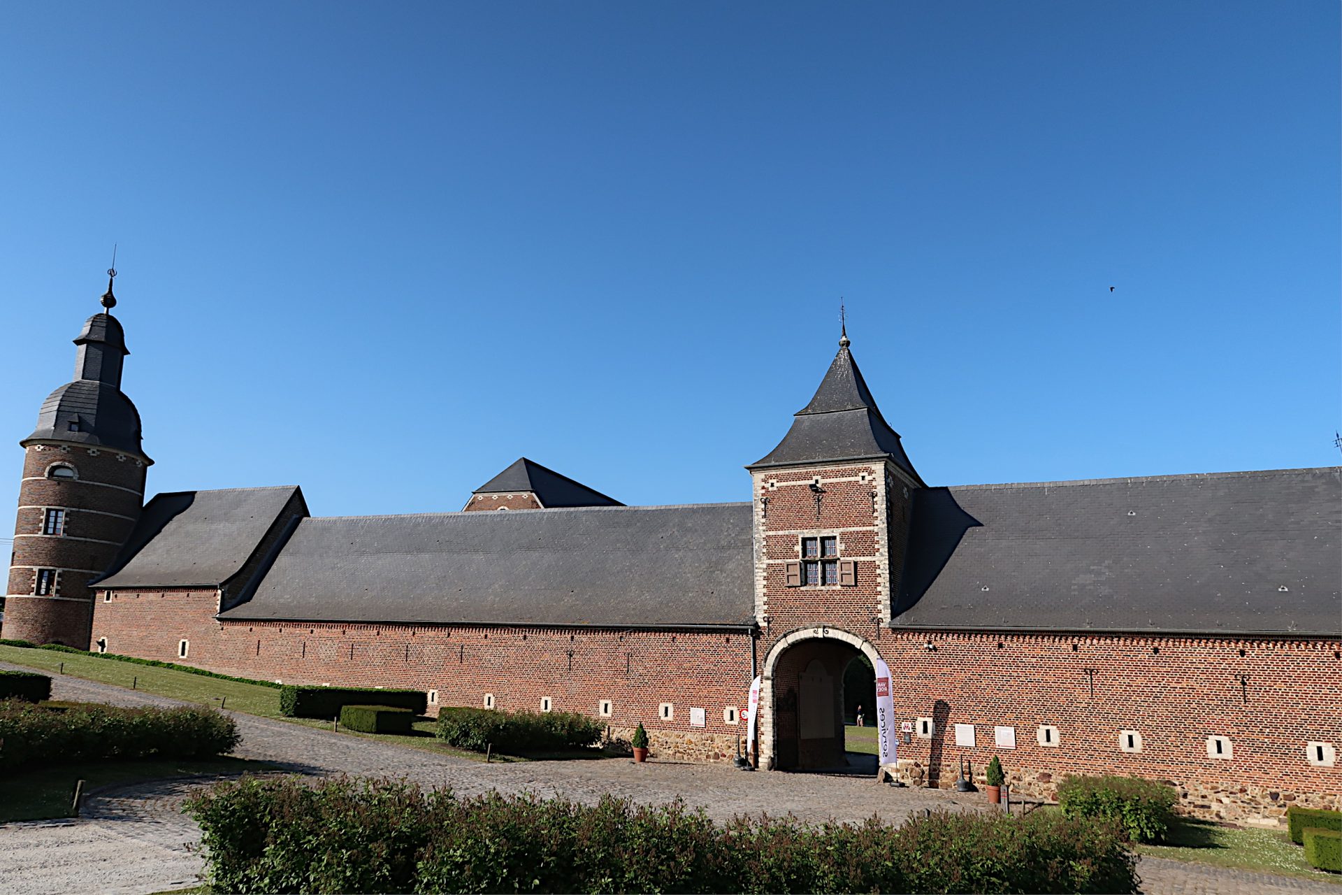 Les longs bâtiments en brique de l’ancienne abbaye de la Ramée à Jauchelette, avec un porche au centre et une tour ronde à gauche, sous un ciel d’un bleu limpide