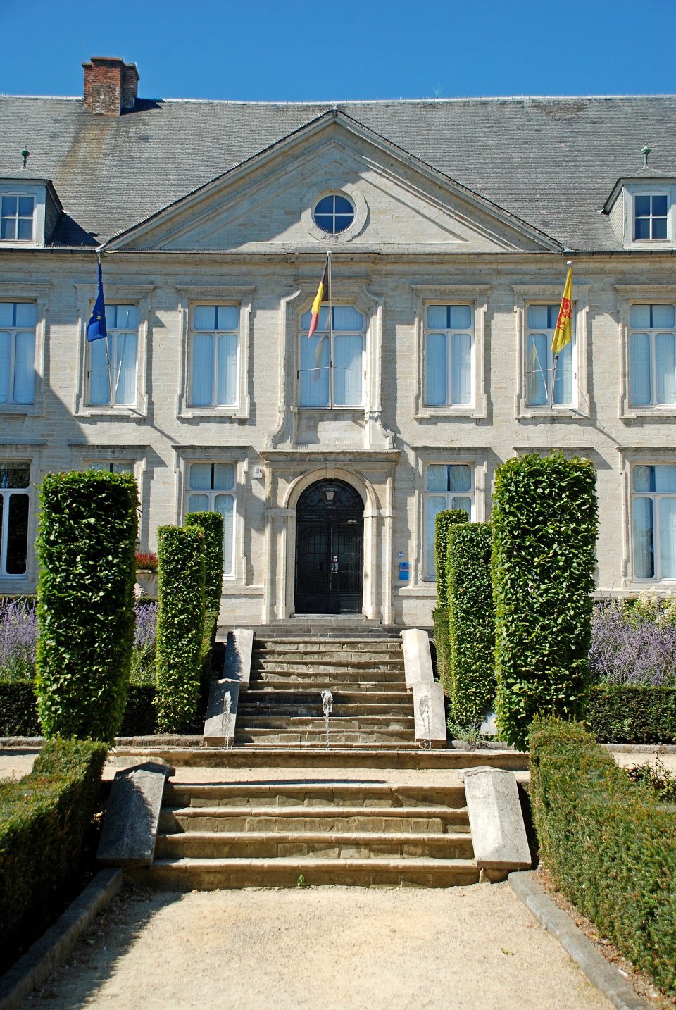La façade en pierre blanche du château Pastur, avec son fronton triangulaire et ses drapeaux, à laquelle mène un escalier de pierre entre de hautes haies taillées