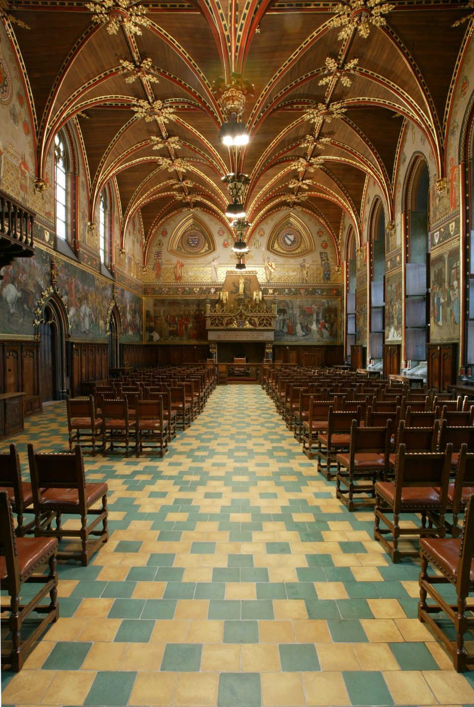 La salle gothique de l’hôtel de ville de Bruges : une voûte en bois peinte aux clés dorées au-dessus de peintures murales, et des rangées de chaises de part et d’autre d’une allée carrelée