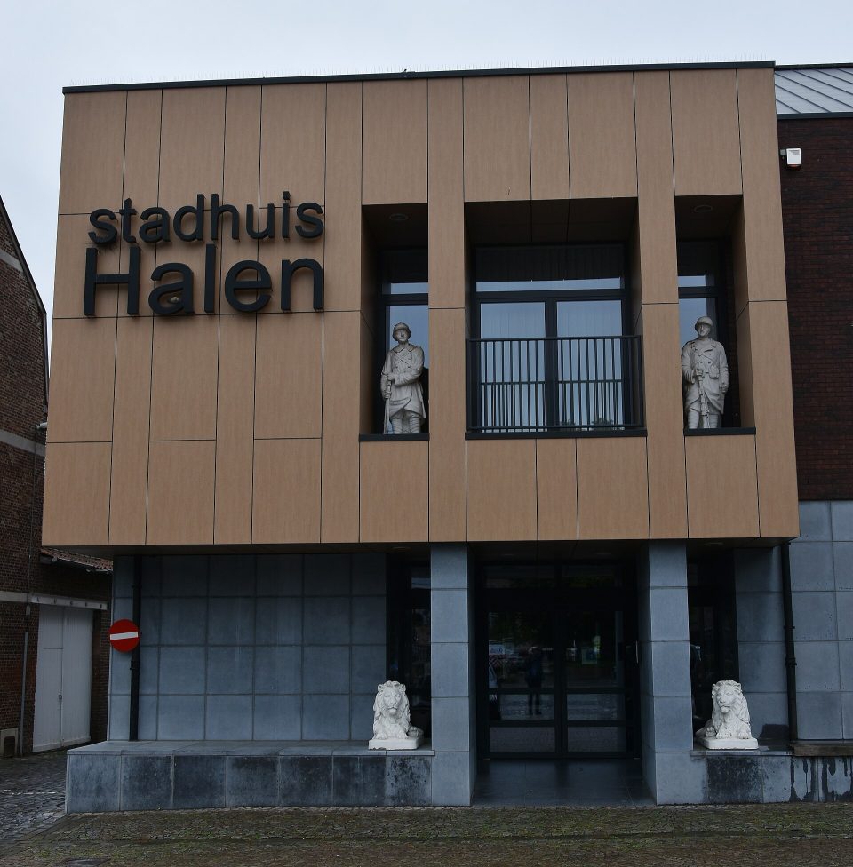 The front of Halen town hall, lettered ‘stadhuis Halen’, with two soldier statues in its windows and two stone lions at the entrance